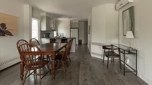 A spacious dining area with a wooden table and chairs, a kitchen in the background, and a modern air conditioning unit on the wall. - Downtown | FREE shuttle to Ski Resort • Ice Rink (Mont-Tremblant)