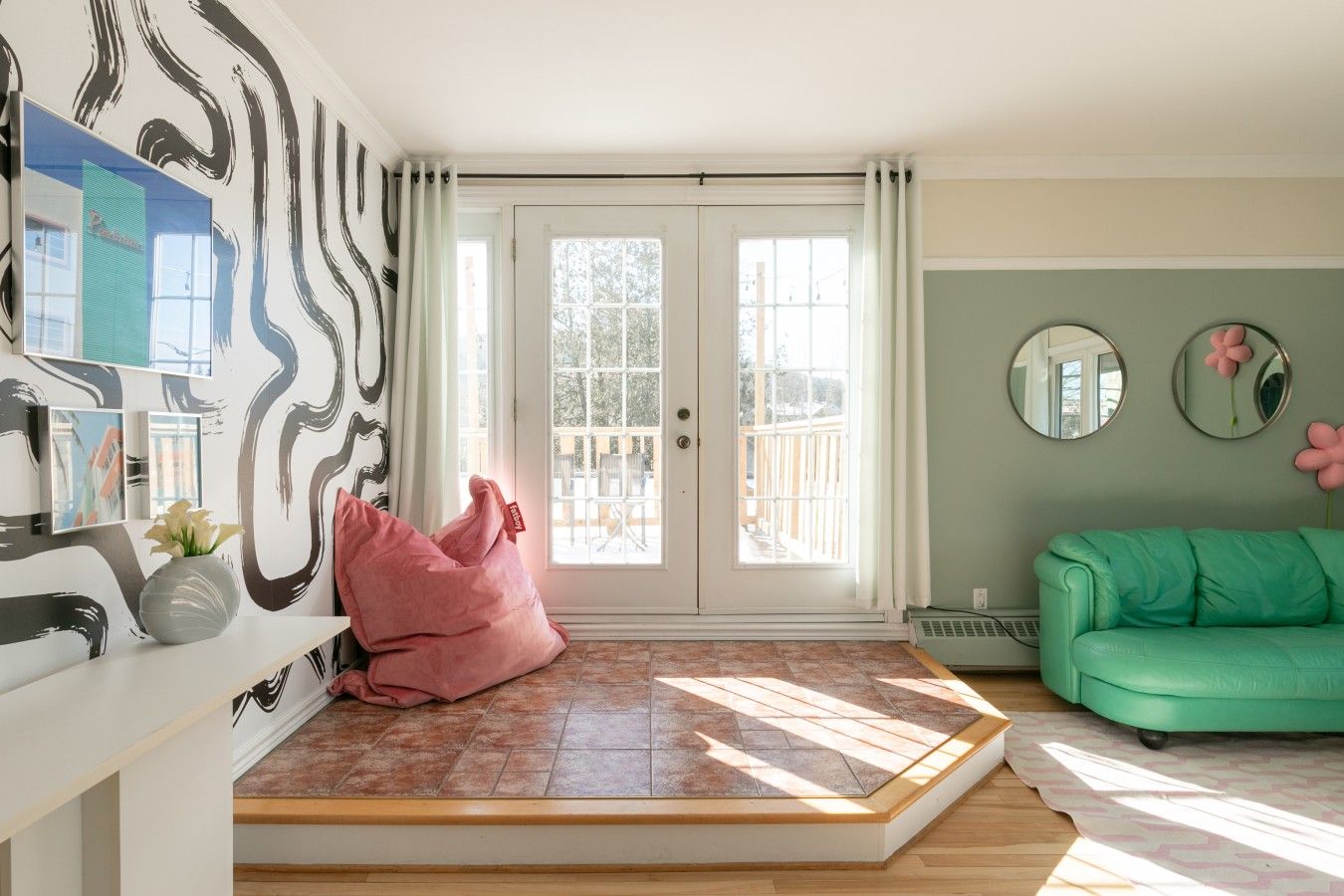 A room with a large window, a green sofa, a pink bean bag, and a wall with abstract art.