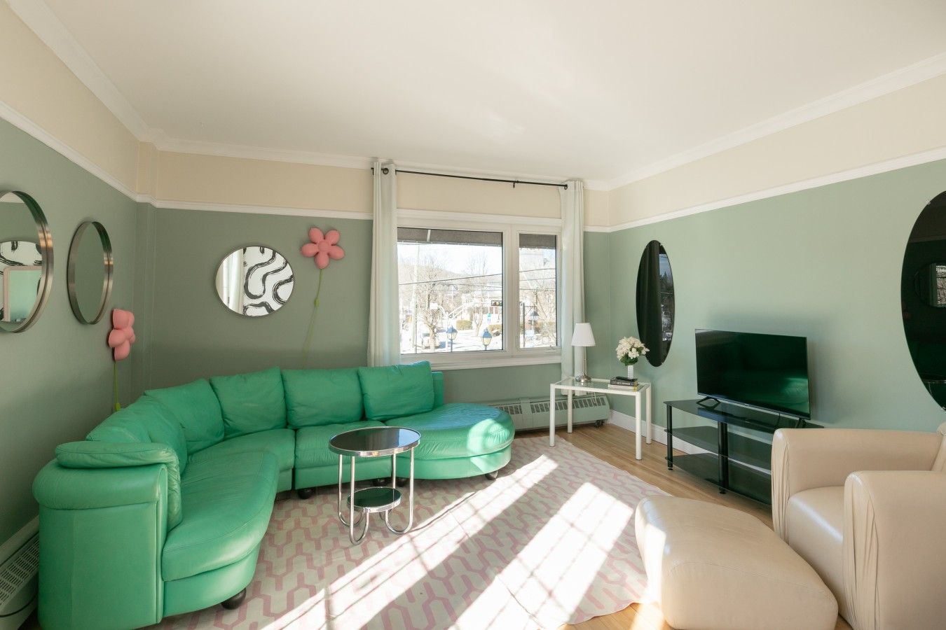 Un salon avec un canapé vert, un fauteuil beige, une petite table ronde, un téléviseur et une fenêtre donnant sur un beau paysage.
