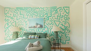 Une chambre avec une couvre-lit verte, deux lampes, un mur à motifs et une serviette pliée sur le lit.