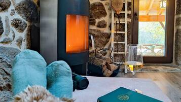 Een knus vertrek met een stenen muur, een houtkachel, een glas wijn en een boek op een tafel.