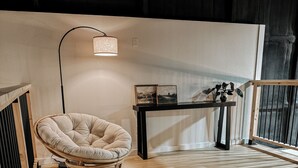 Una habitación acogedora con piso de madera, un sillón con cojines, una mesita con fotografías, una lámpara de pie y una barandilla.