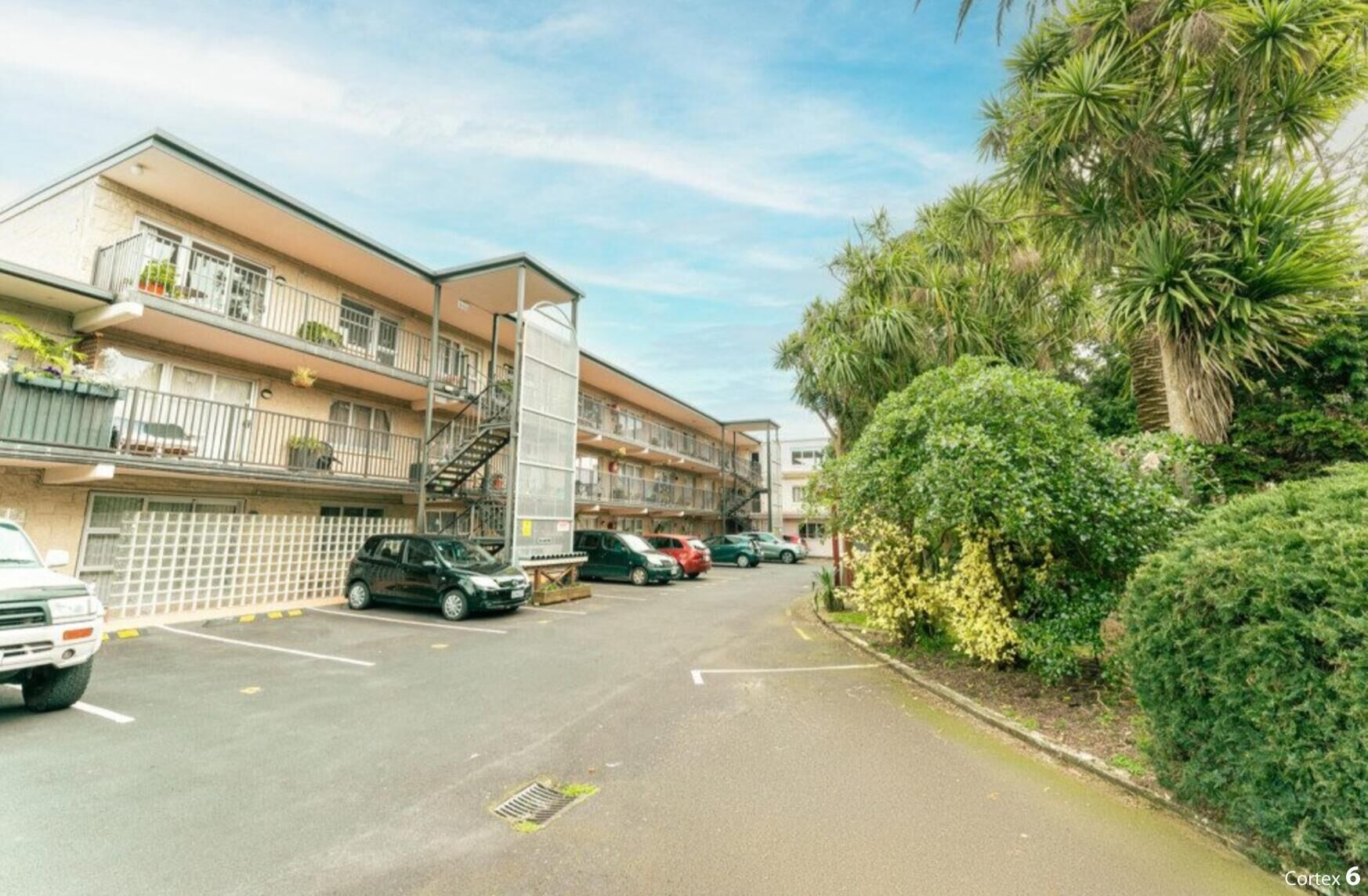 Un edificio residencial de varios pisos con balcones, una zona de estacionamiento y abundante vegetación.