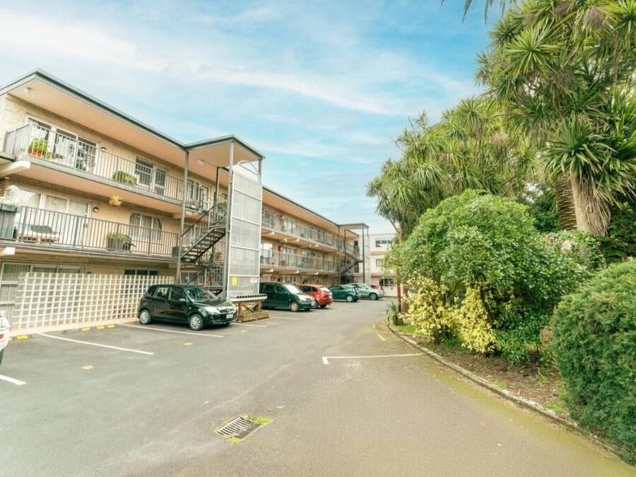Un edificio residencial de varios pisos con balcones, una zona de estacionamiento y abundante vegetación.