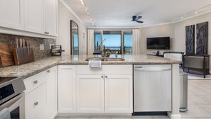 A modern kitchen with white cabinets, granite countertops, and a large island. There is a view of the ocean through large windows.