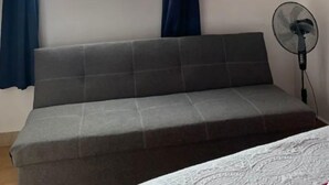 Une pièce avec un canapé gris, un téléviseur fixé au mur, une fenêtre avec des rideaux bleus et un ventilateur.