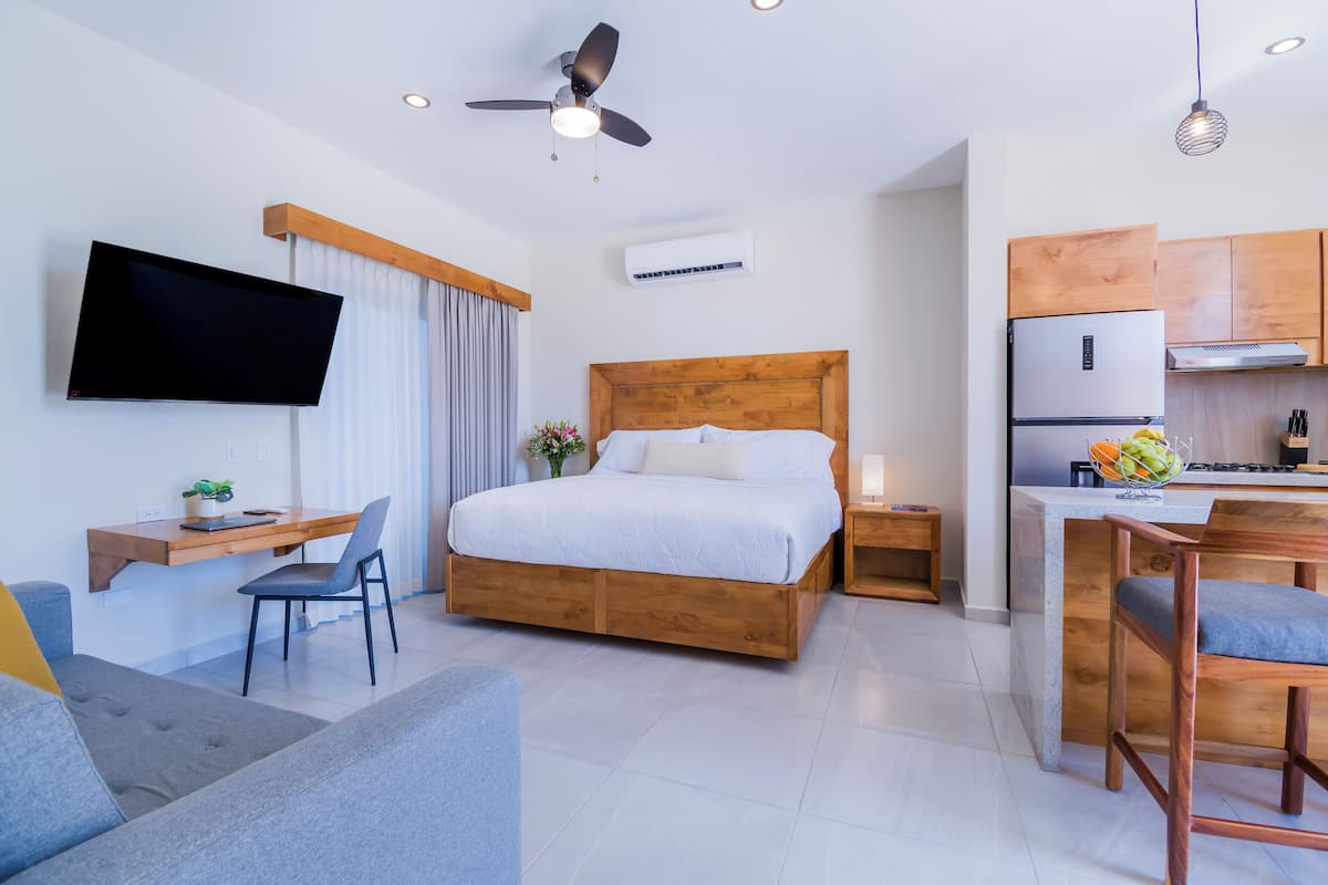 A modern hotel room with a wooden bed, a desk, a flat-screen TV, a dining area, and a kitchenette.
