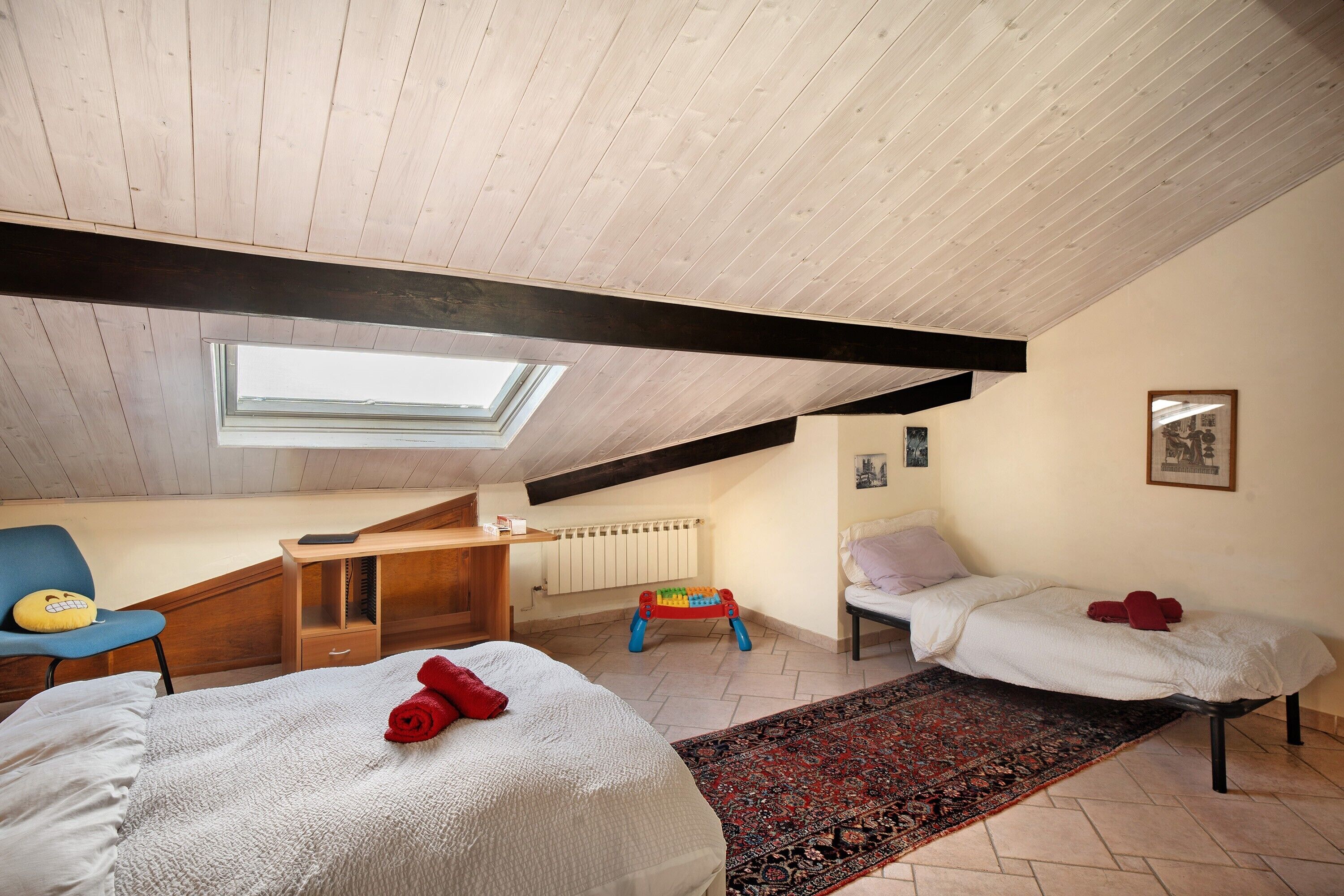 A room with a sloped ceiling, a skylight, a bed with a white cover, a small desk, a chair, a colorful play area, and a bed with a red pillow.