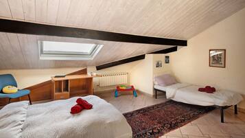A room with a sloped ceiling, a skylight, a bed with a white cover, a small desk, a chair, a colorful play area, and a bed with a red pillow.
