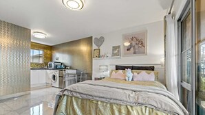 A modern bedroom with a large bed, a washing machine, and a kitchen area. - 11 of 43 woodward road mount albert (Auckland)