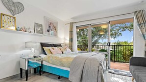 A bedroom with a bed, bedside table, lamp, and a view of the outdoors. - 11 of 43 woodward road mount albert (Auckland)