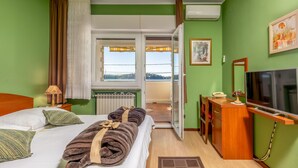 Ein Hotelzimmer mit Bett, Schreibtisch mit Telefon, Fernseher und Blick auf einen Balkon mit Geländer.