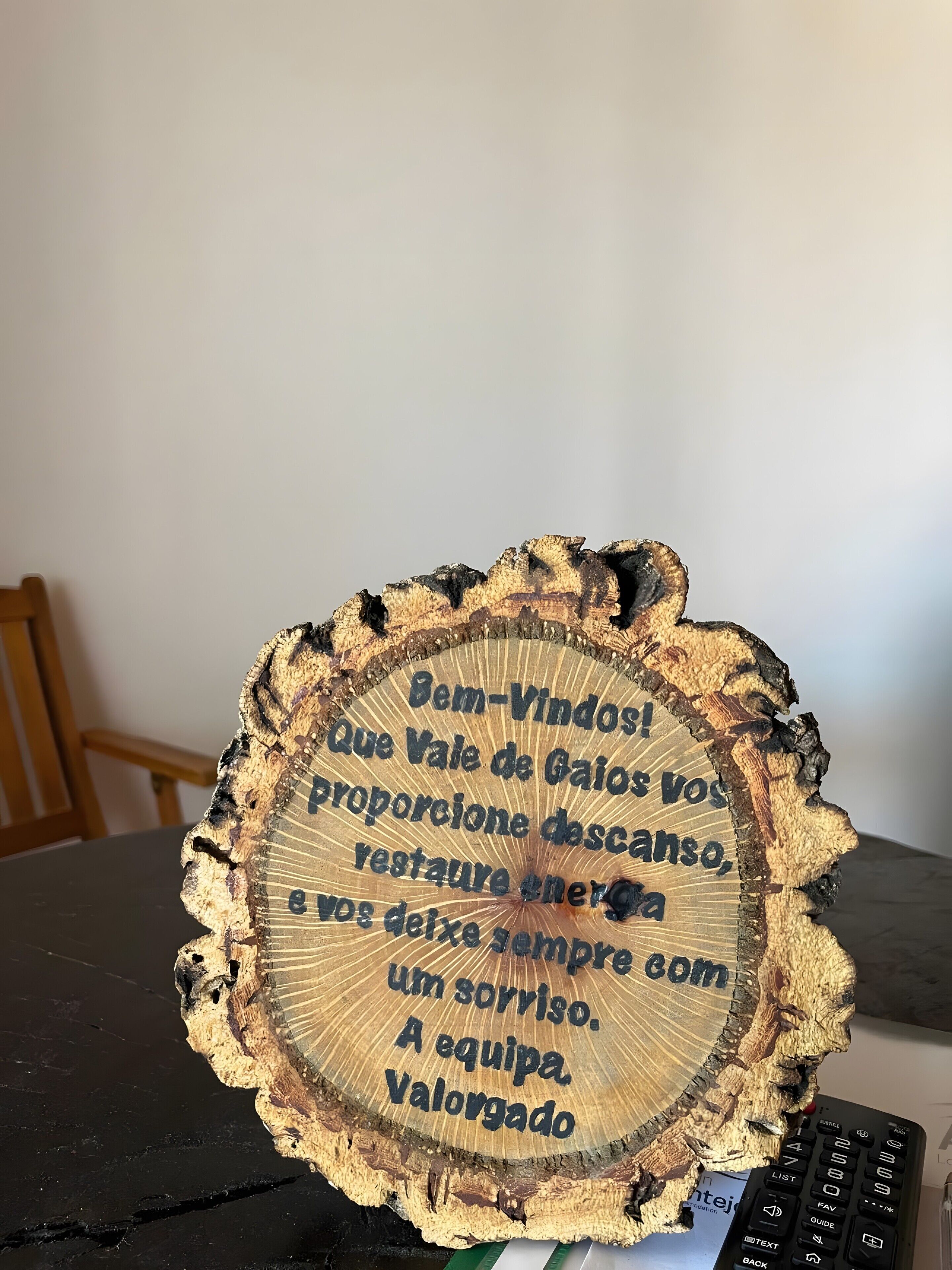A wooden coaster with text in Portuguese, placed on a table with a keyboard and a chair in the background.