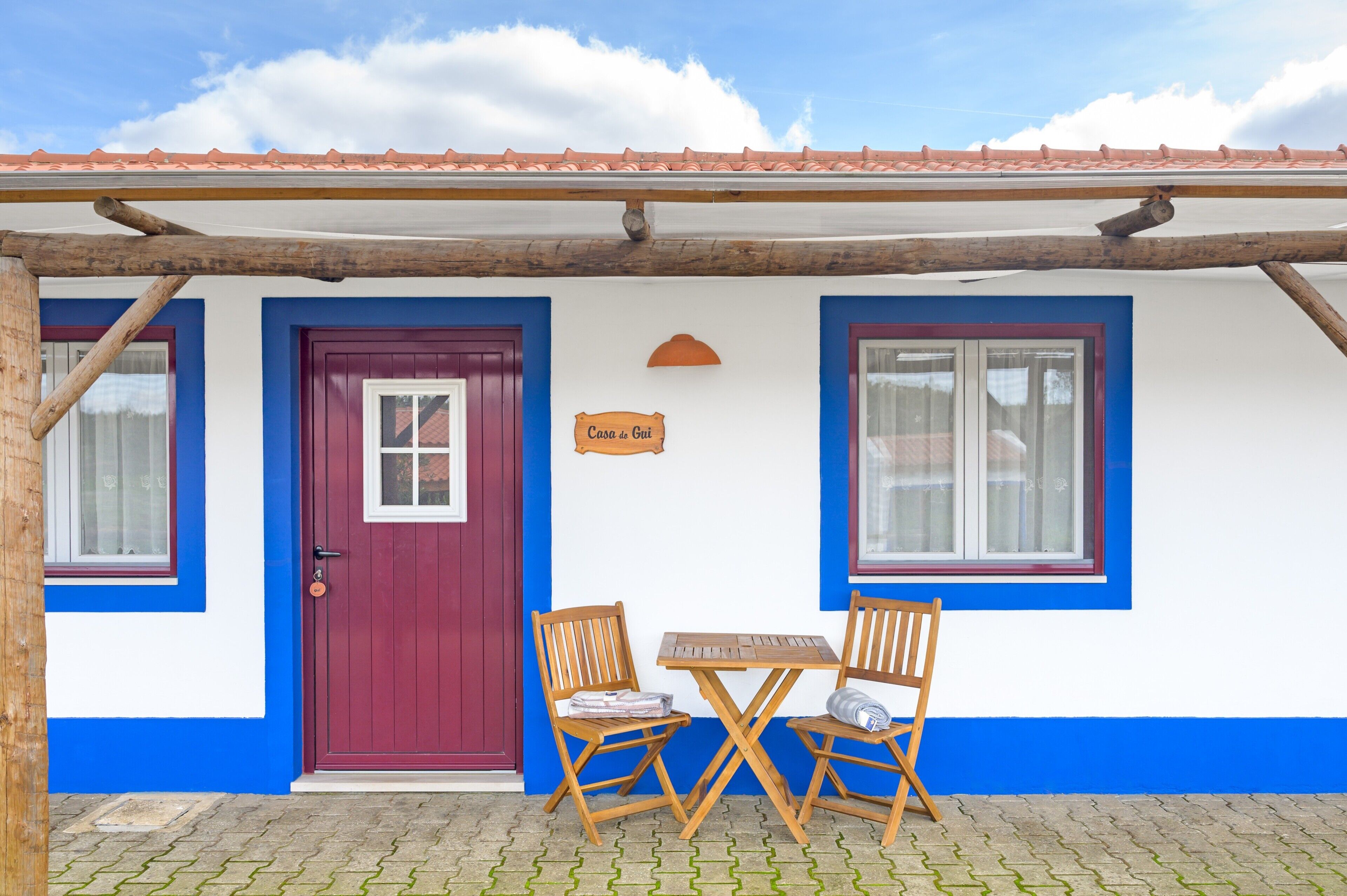 A building with a red door, blue trim, and a sign reading 'Casa de Gui.' There are two wooden chairs and a table outside.