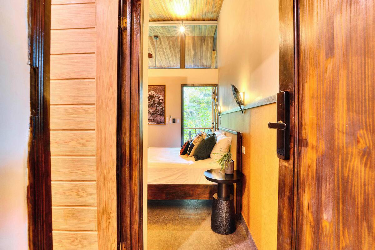 Eine Holztür führt in ein Schlafzimmer mit einem Bett, einem Nachttisch und einem Fenster mit Blick auf Grünflächen.