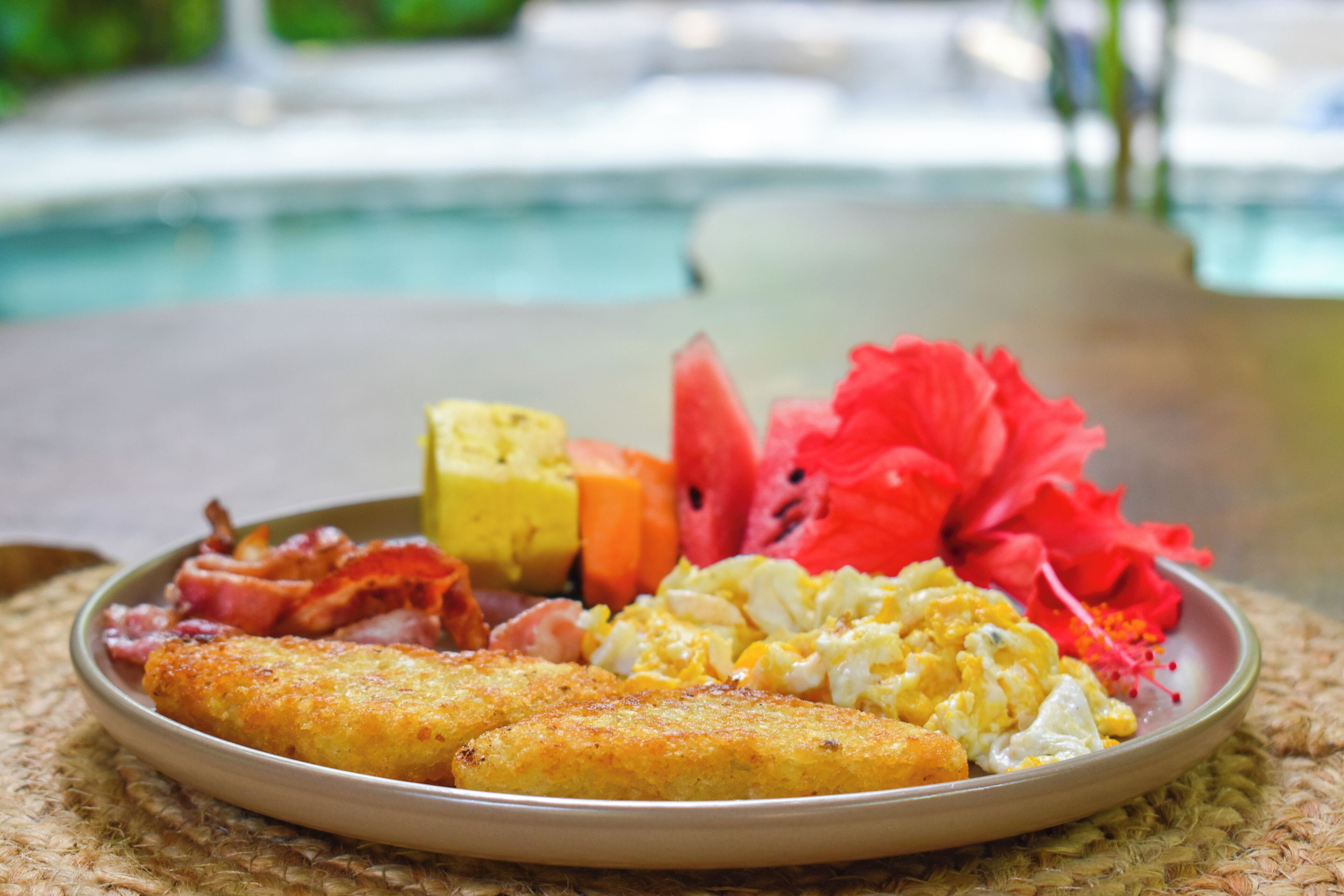 A plate of breakfast food including eggs, bacon, and fruit, with a pool in the background.