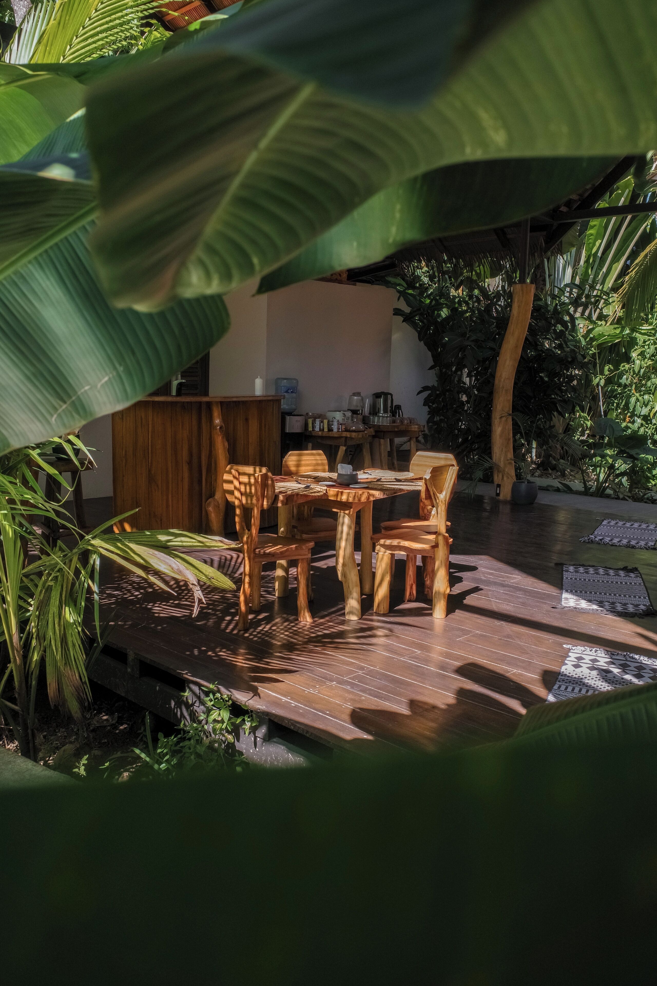 A wooden outdoor dining area with a table and chairs, surrounded by lush greenery and a water feature.
