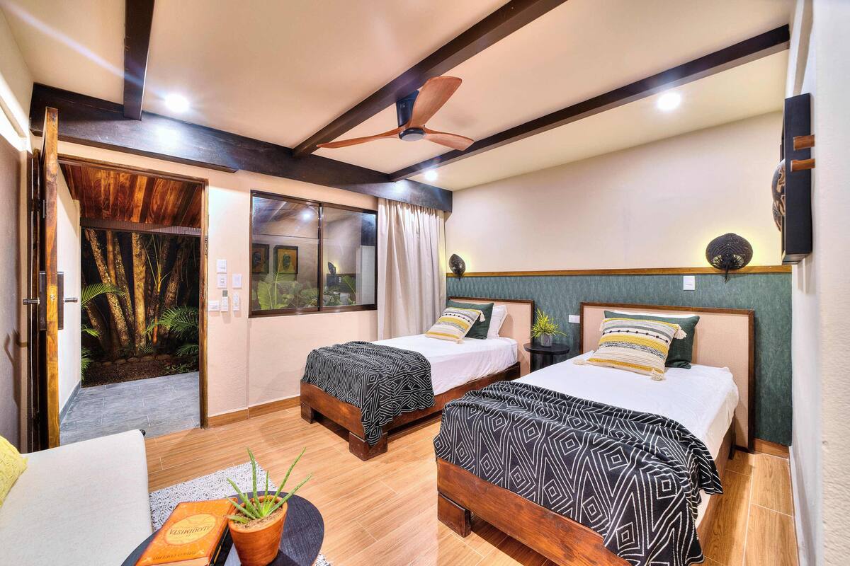 Ein Hotelzimmer mit zwei Betten, einem Deckenventilator, einem kleinen Tisch mit einer Pflanze und Blick auf einen Wald durch eine Glastür.