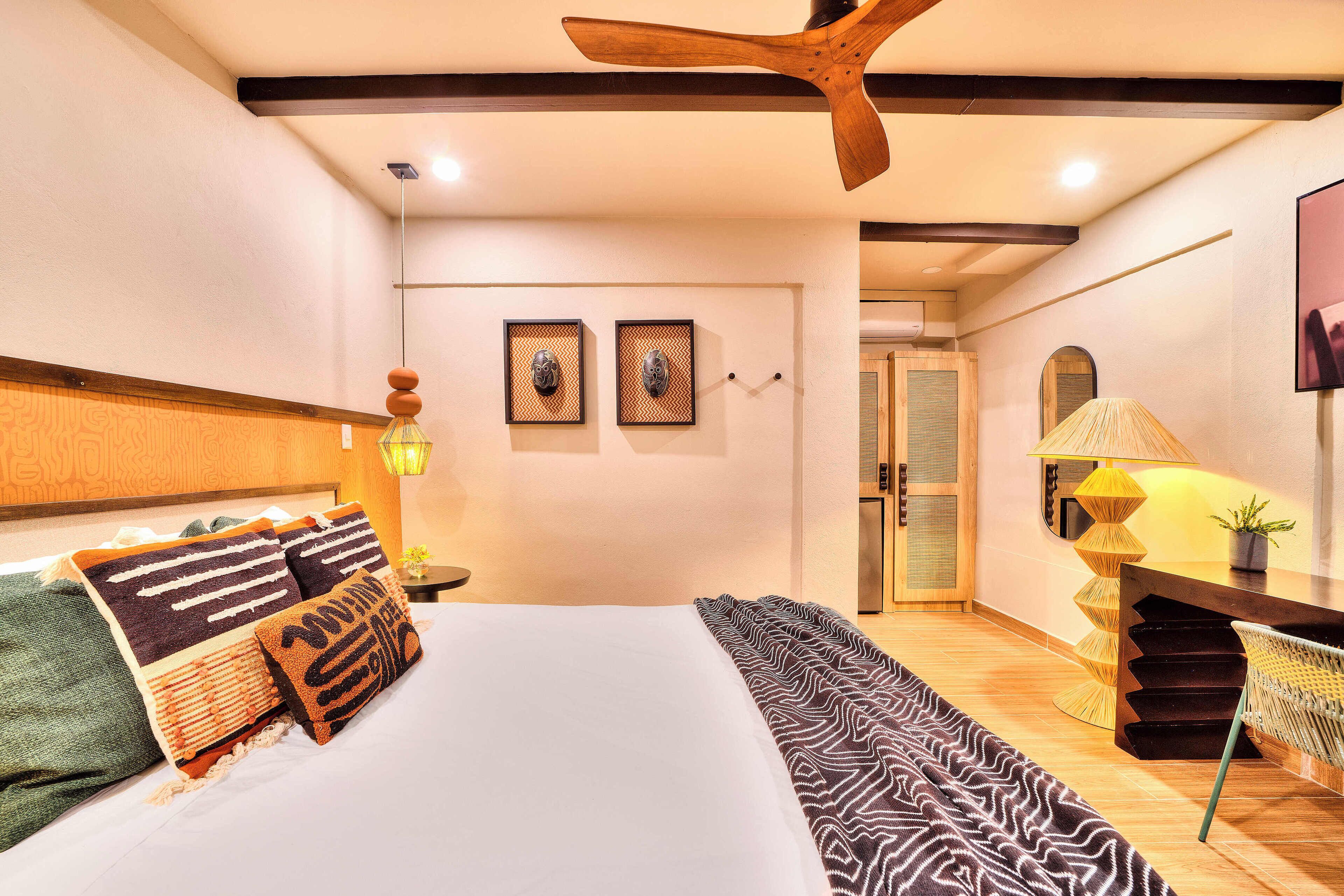 A bedroom with a large bed, wooden ceiling fan, two framed pictures on the wall, and a desk with a lamp.