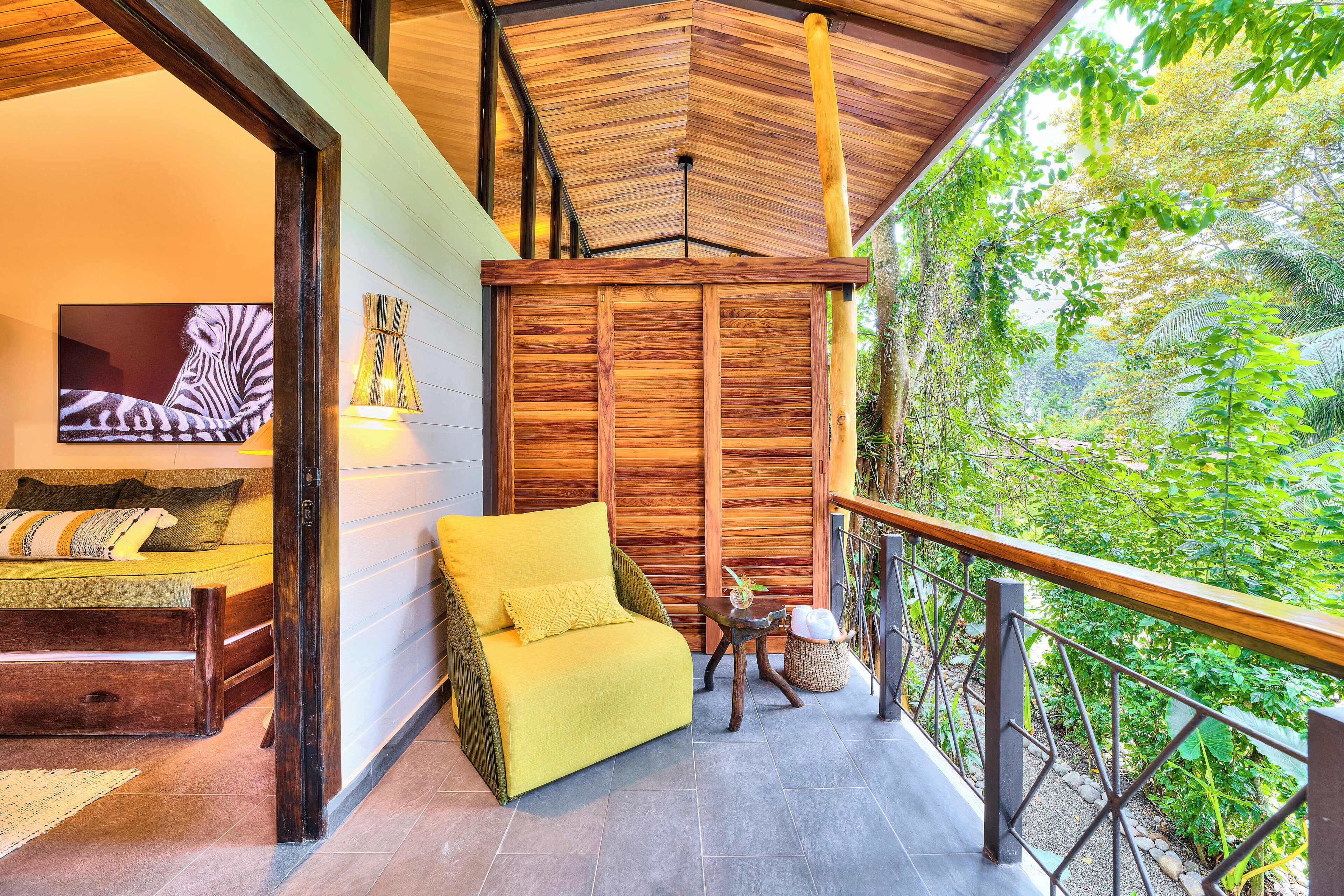 Un balcón con baranda de madera, un sofá amarillo, una mesita con lámpara y vista a vegetación exuberante.