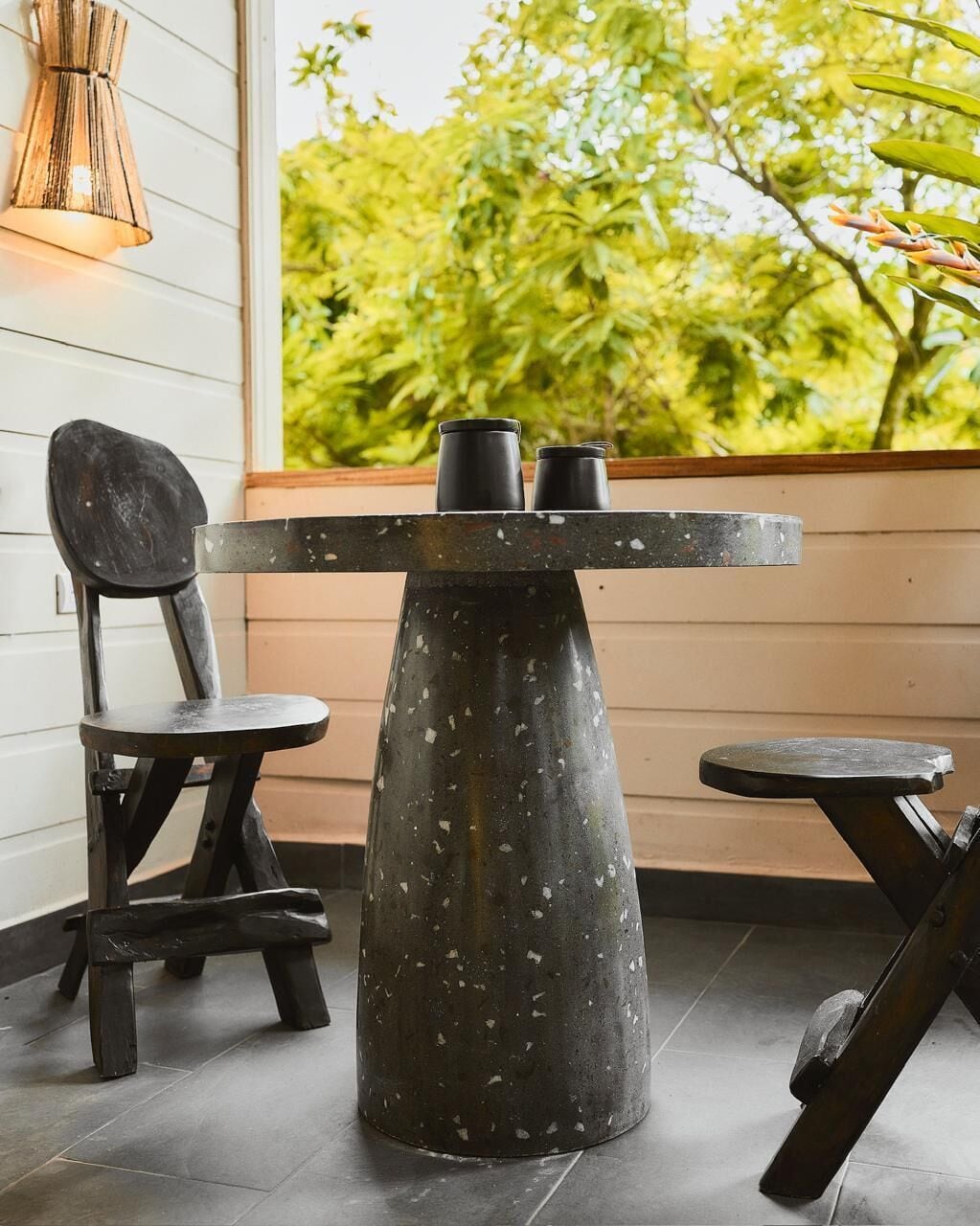 A table with two black chairs, a pendant light, and a view of greenery outside.