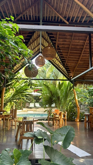 Relax in a spacious room with high ceilings, wooden tables, and lush green plants, perfect for socializing and taking in the serene ambiance.