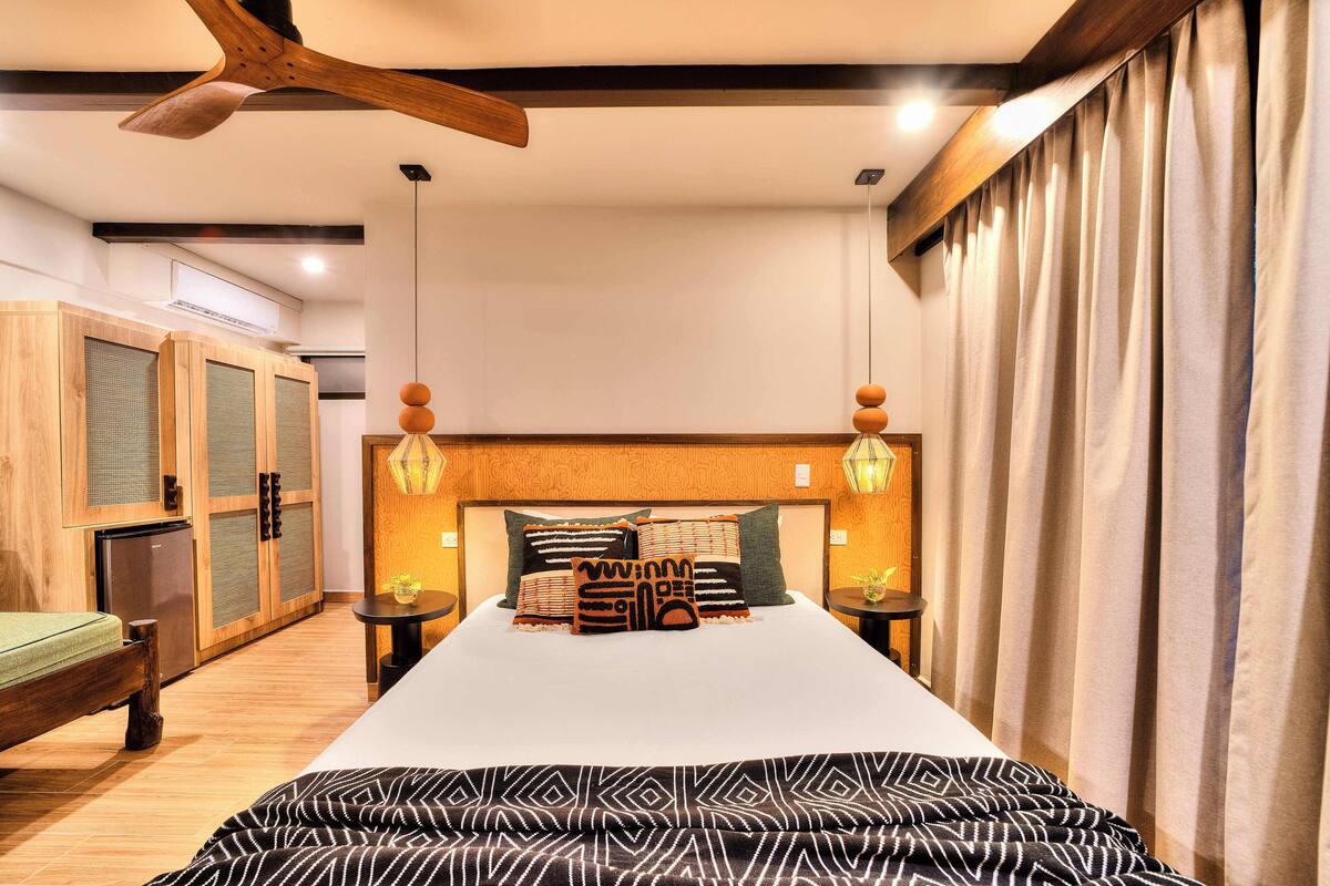 Ein Hotelzimmer mit einem großen Bett, einem Holzkopfbrett und einem Deckenventilator. In der Ecke steht ein kleiner Tisch mit einer Lampe und eine Bank.