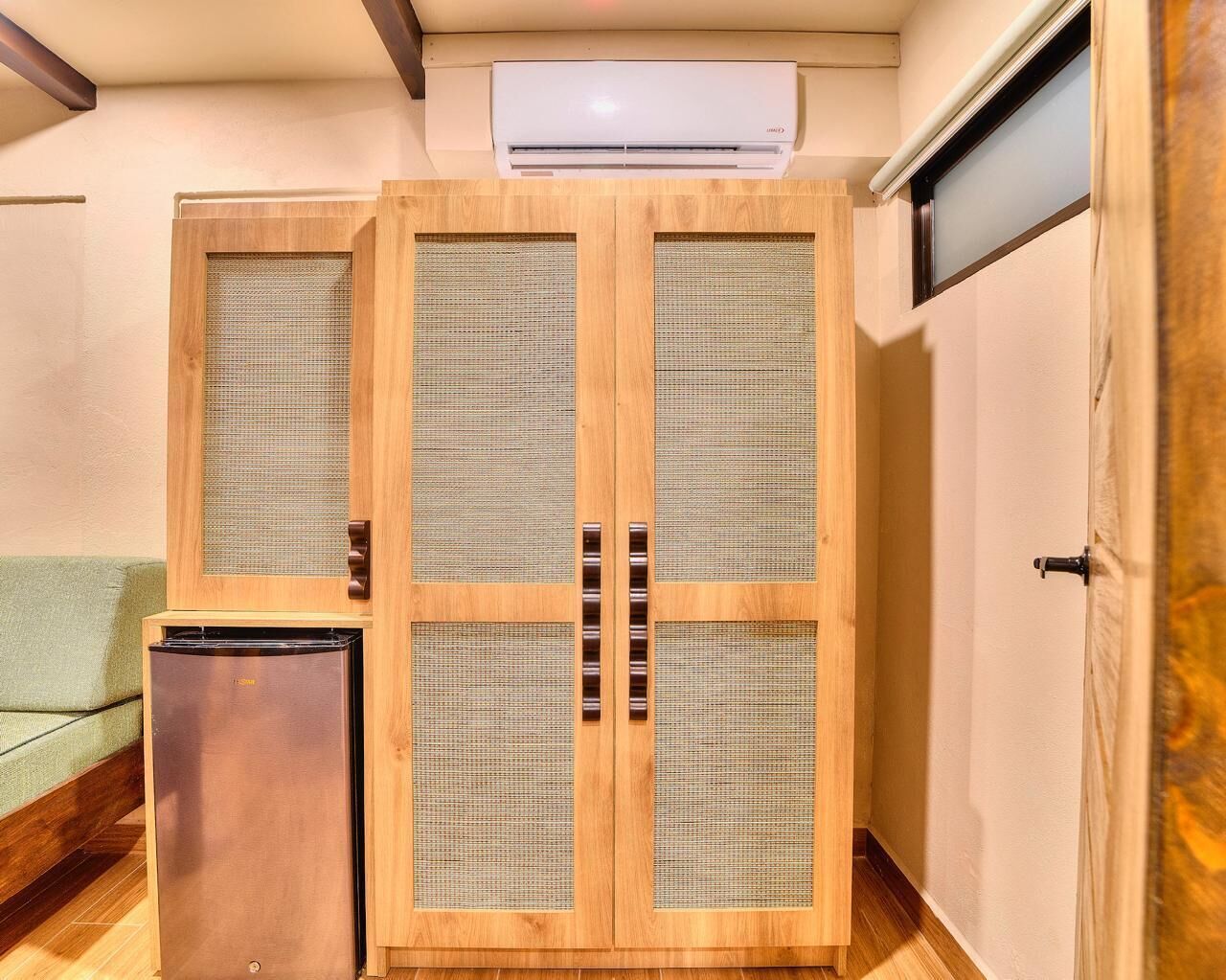 A room with a wooden wardrobe, a small refrigerator, and an air conditioning unit on the ceiling.