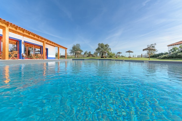 A swimming pool with clear blue water, wooden deck chairs, and a covered seating area.