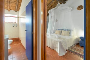 Une chambre avec un lit recouvert d’une moustiquaire, une salle de bain avec des toilettes et un lavabo, et une porte bleue.