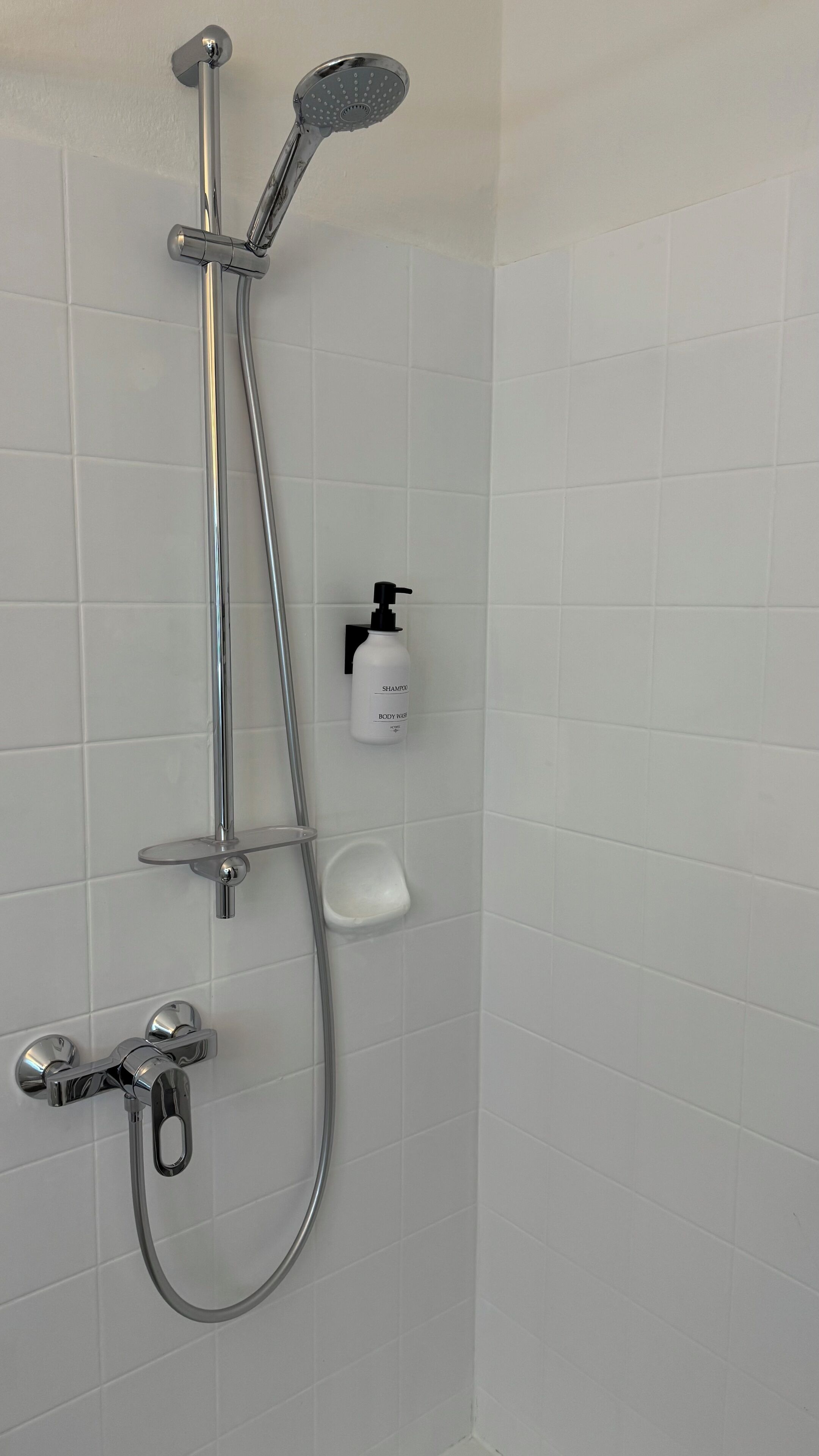 Een douche met een handdouche, een wandmengkraan en een handdouche. Er is een shampoo flesje aan de muur zichtbaar.