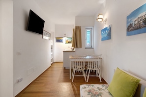 Een smalle gang met een eetgedeelte, inclusief een witte tafel met stoelen, een aan de muur bevestigde televisie en een raam met gordijnen.