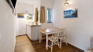 Une petite pièce avec une table et des chaises en bois, une fenêtre avec des rideaux, un téléviseur fixé au mur et des tableaux encadrés.