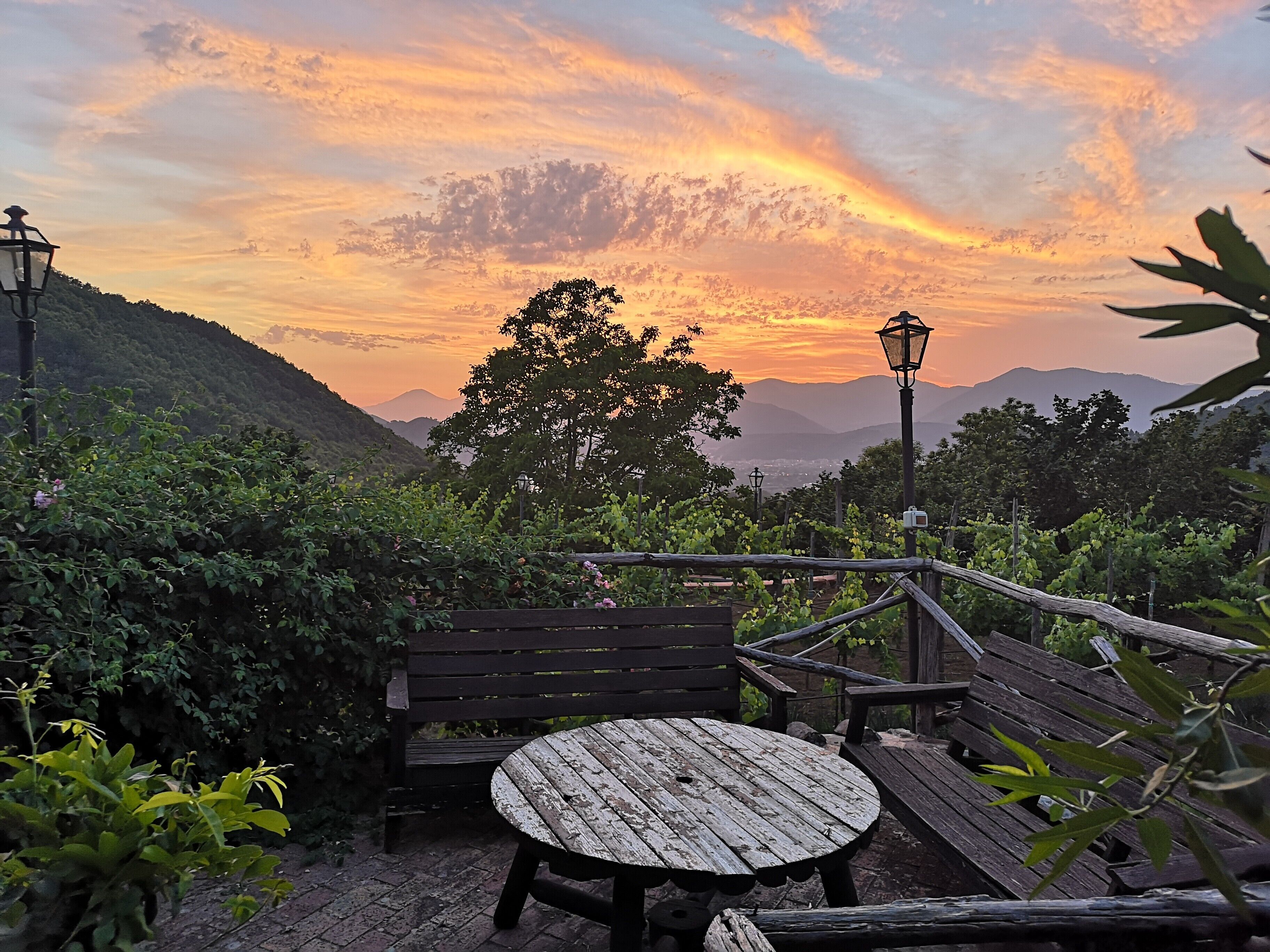 A wooden bench and table set on a patio with a view of a sunset over a mountainous landscape.