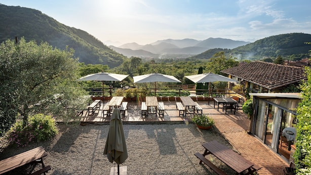 A patio with wooden tables and umbrellas, surrounded by greenery and overlooking a valley.