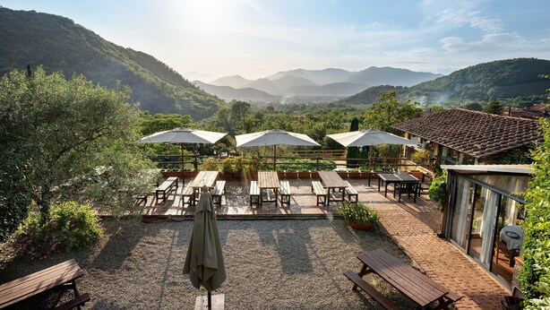 A patio with wooden tables and umbrellas, surrounded by greenery and overlooking a valley.
