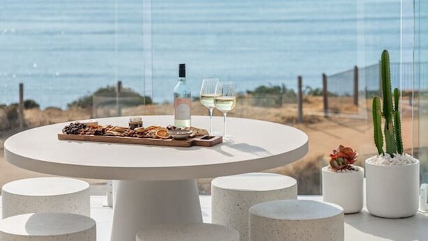 A round table with a bottle and glasses, surrounded by concrete stools, overlooking a beach with a fence and cacti. - Sunsets ~ Rooftop, Incredible views, 4 KING rooms (Aldinga Beach)