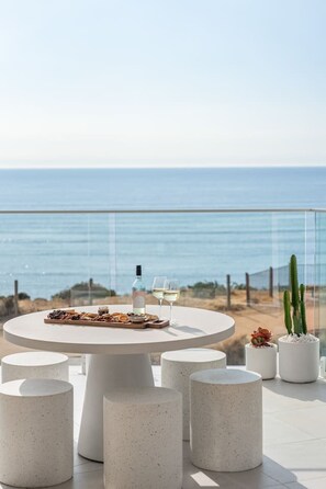 A round table with a bottle and glasses, surrounded by concrete stools, overlooking a beach with a fence and cacti. - Sunsets ~ Rooftop, Incredible views, 4 KING rooms (Aldinga Beach)