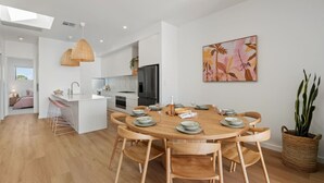 A modern kitchen with white cabinetry, a central island, and a dining area with a wooden table and chairs. - Sunsets ~ Rooftop, Incredible views, 4 KING rooms (Aldinga Beach)