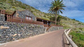 Edifici de pared de pedra amb una tanca de fusta, un camí corbat i una palmera.