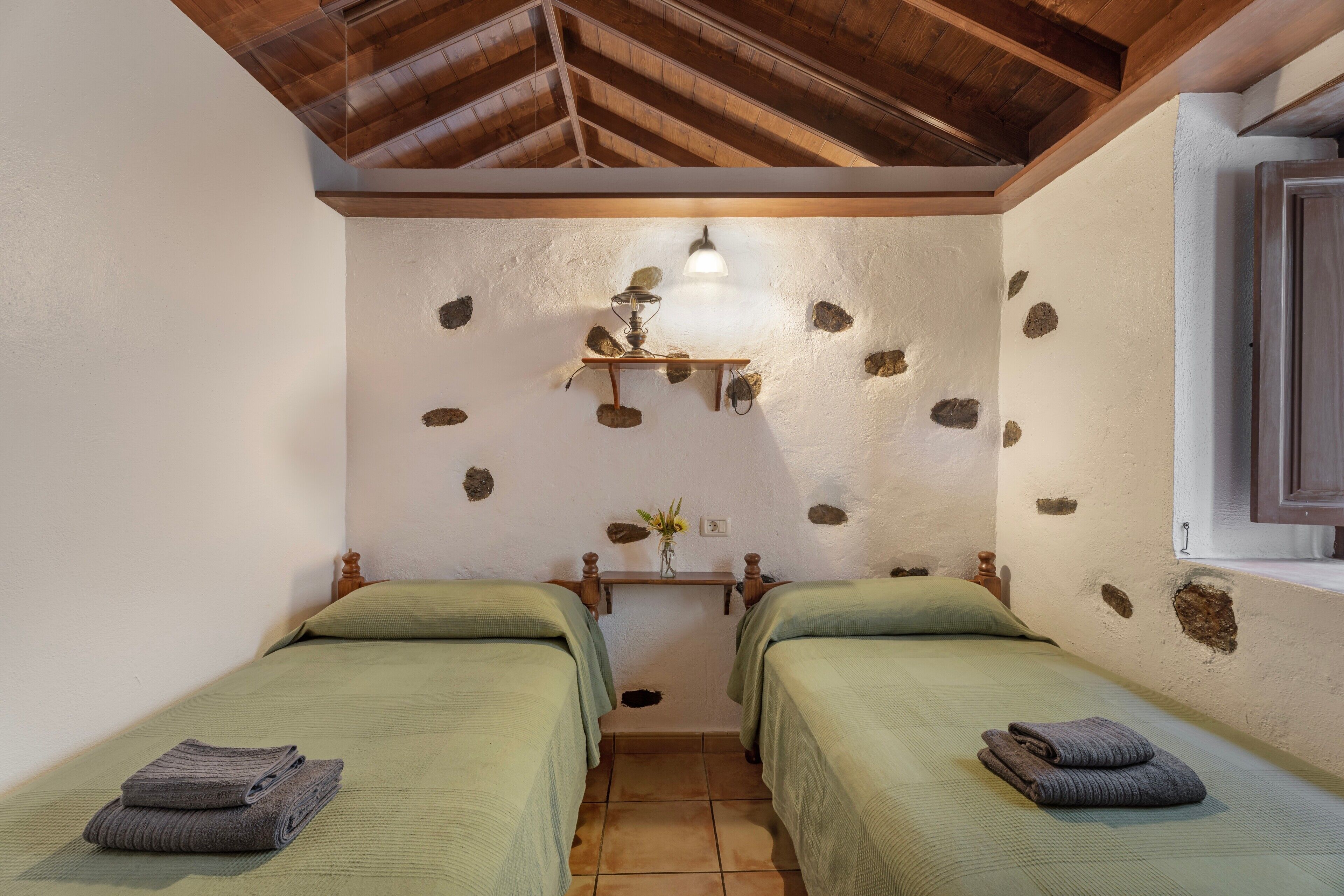 Una habitación con dos camas, techo de madera, pared de piedra, una estantería con una lámpara y una ventana.