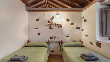 Una habitación con dos camas, techo de madera, pared de piedra, una estantería con una lámpara y una ventana.