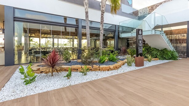 A modern building with a glass facade, outdoor seating area, and a landscaped garden with various plants.