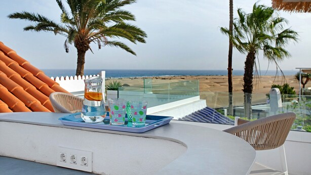 A balcony with a table, chairs, a tray with glasses and a bottle, and a view of the beach and palm trees.