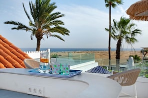Un balcón con mesa, sillas, una bandeja con vasos y una botella, y vista a la playa y palmeras.