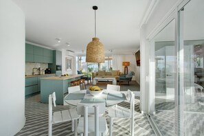 Una cucina moderna con mobili color petrolio, una zona pranzo con tavolo in vetro e un soggiorno con divano e tavolino.