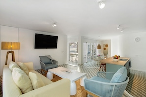 Een moderne woonkamer met een bank, fauteuil en salontafel, een flatscreen-tv, en een keukenhoek met een eettafel en bar.'