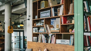 A cozy café with wooden shelves filled with books and magazines, a wooden bench with patterned cushions, and a guitar hanging on the wall. - Domus Tlv (Tel Aviv)