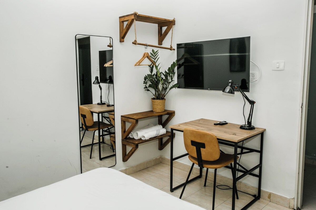 A small, modern room with a wooden desk, a chair, a mirror, a potted plant, and a TV mounted on the wall.