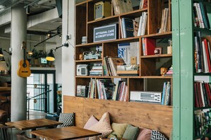 Un café chaleureux avec des étagères en bois remplies de livres et de magazines, un banc en bois agrémenté de coussins à motifs et une guitare accrochée au mur.